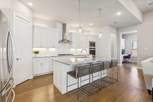 kitchen with stainless steel appliances, white cabinetry, a kitchen island with sink, and wall chimney range hood