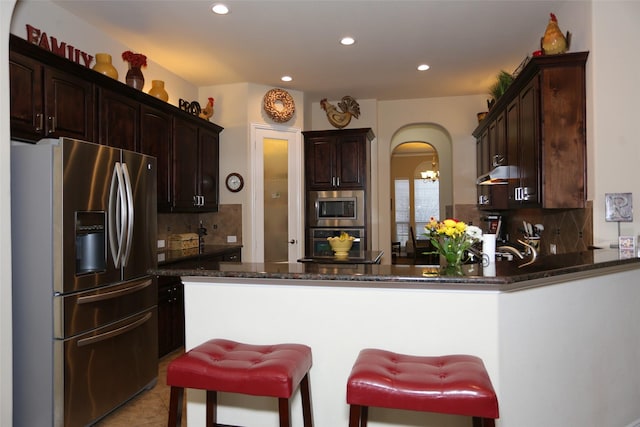 kitchen featuring dark stone counters, decorative backsplash, dark brown cabinets, kitchen peninsula, and stainless steel appliances