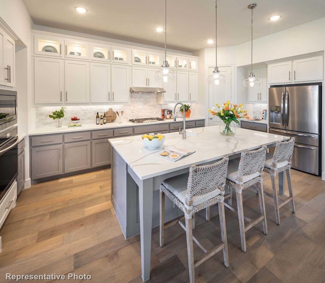 kitchen with appliances with stainless steel finishes, pendant lighting, a kitchen breakfast bar, light stone countertops, and a center island with sink