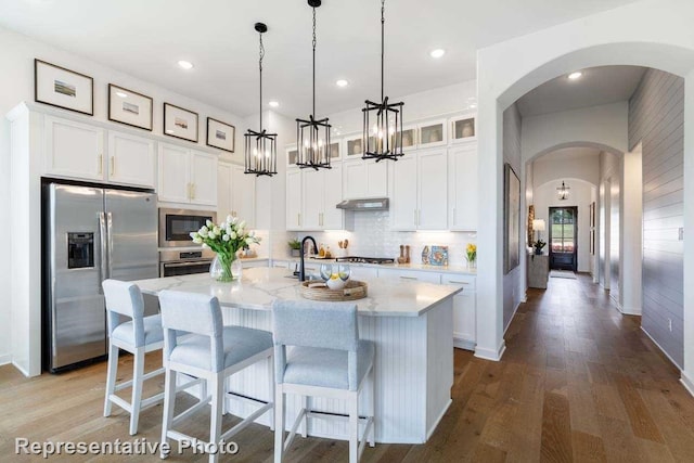 kitchen with white cabinets, light stone countertops, an island with sink, decorative light fixtures, and stainless steel appliances