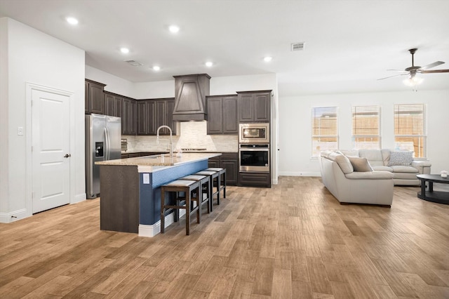 kitchen with stainless steel appliances, light hardwood / wood-style flooring, a kitchen bar, a center island with sink, and custom exhaust hood