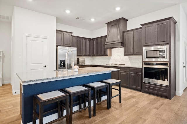 kitchen with appliances with stainless steel finishes, dark brown cabinetry, a center island with sink, and custom range hood