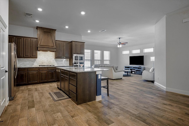 kitchen featuring premium range hood, stainless steel appliances, ceiling fan, a breakfast bar area, and an island with sink