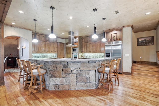 kitchen featuring decorative backsplash, light wood-type flooring, wall chimney exhaust hood, built in appliances, and decorative light fixtures