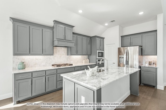 kitchen with gray cabinetry, custom exhaust hood, a kitchen island with sink, appliances with stainless steel finishes, and light stone counters