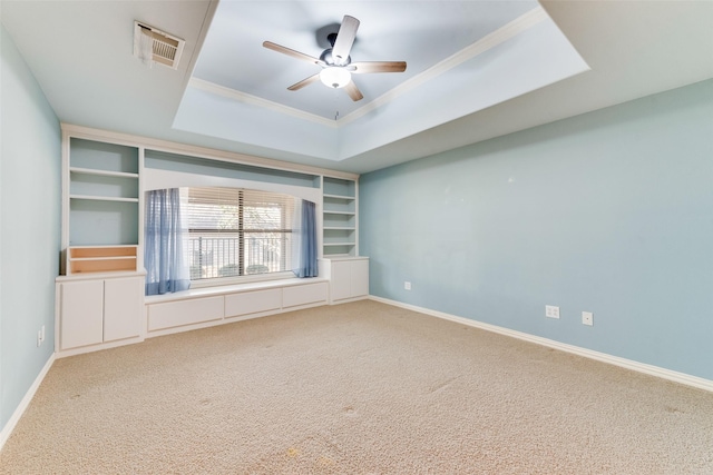 empty room with a raised ceiling, crown molding, ceiling fan, built in shelves, and carpet floors