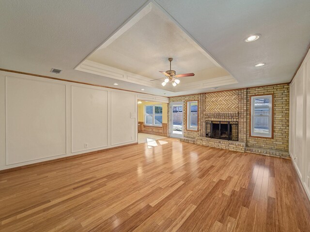 unfurnished living room featuring a raised ceiling, ceiling fan, a brick fireplace, and light wood-type flooring