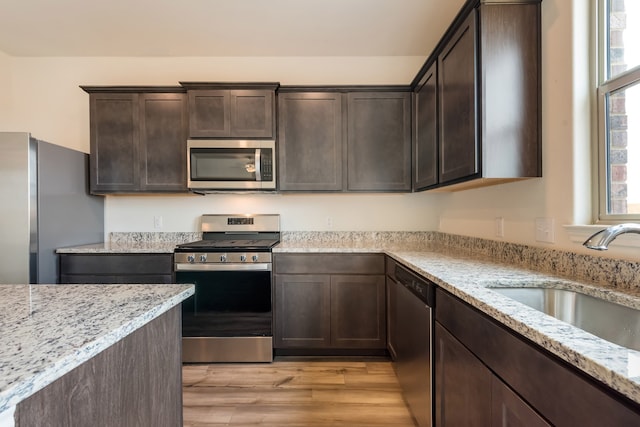 kitchen with appliances with stainless steel finishes, light stone counters, dark brown cabinets, sink, and light hardwood / wood-style flooring