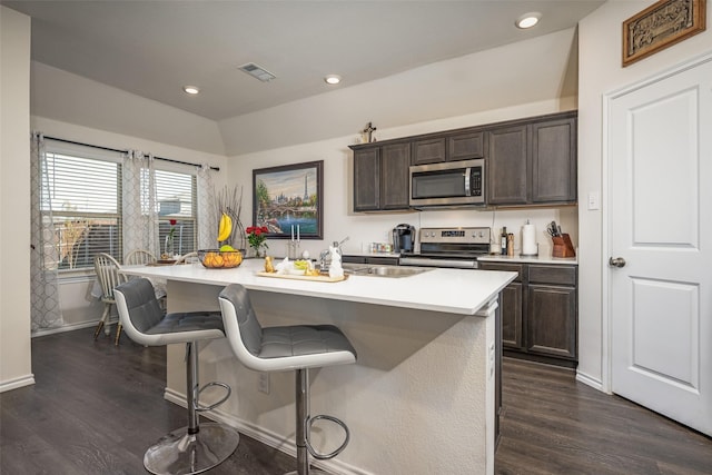 kitchen with dark brown cabinetry, stainless steel appliances, dark wood-type flooring, a kitchen bar, and a center island with sink