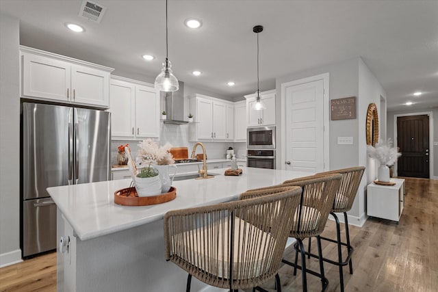 kitchen with a center island with sink, hanging light fixtures, stainless steel appliances, white cabinets, and wall chimney exhaust hood