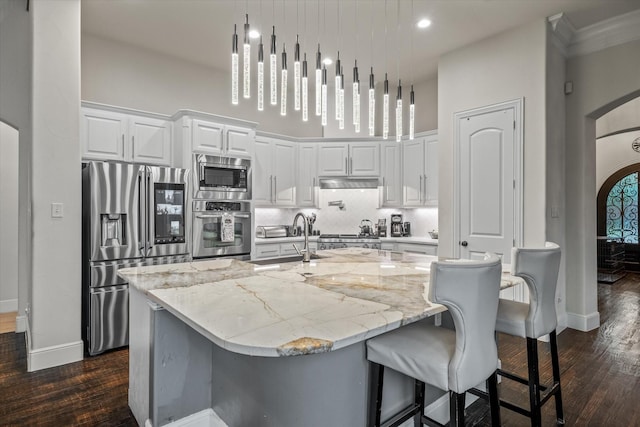 kitchen with light stone countertops, stainless steel appliances, a kitchen island with sink, decorative light fixtures, and white cabinetry