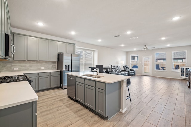 kitchen with sink, gray cabinets, an island with sink, stainless steel appliances, and decorative backsplash