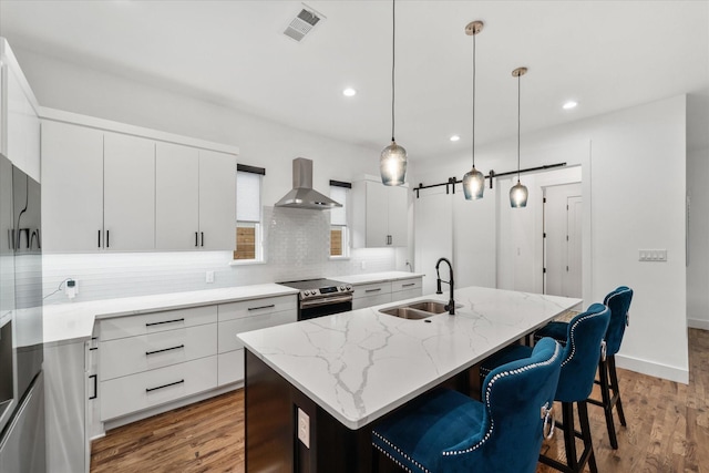 kitchen with sink, wall chimney exhaust hood, stainless steel range with electric cooktop, a kitchen island with sink, and white cabinets