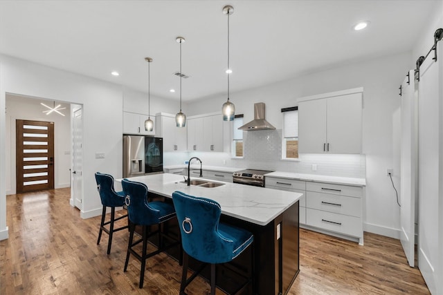 kitchen featuring a kitchen island with sink, wall chimney range hood, sink, a barn door, and stainless steel appliances