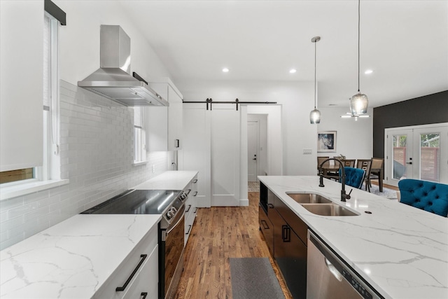 kitchen featuring sink, wall chimney range hood, a barn door, decorative light fixtures, and appliances with stainless steel finishes