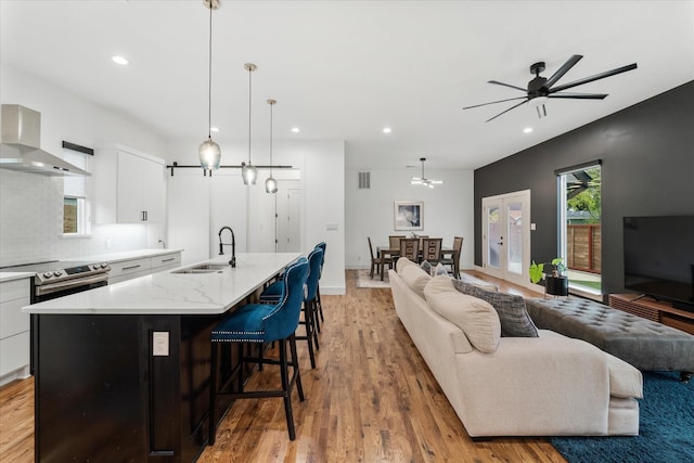 kitchen featuring white cabinetry, sink, wall chimney range hood, an island with sink, and decorative light fixtures