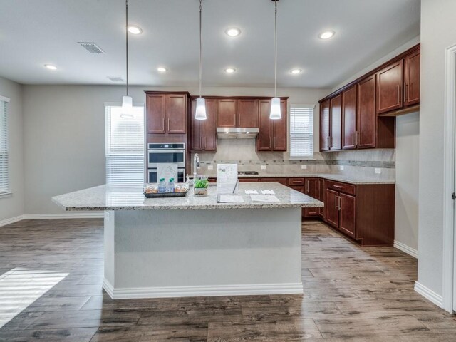 kitchen with light stone countertops, backsplash, a kitchen island with sink, pendant lighting, and hardwood / wood-style flooring