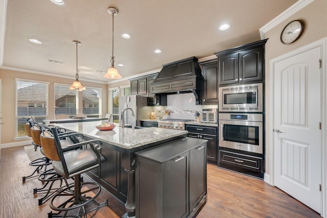 kitchen with a breakfast bar area, stainless steel appliances, an island with sink, decorative light fixtures, and custom exhaust hood