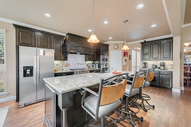 kitchen with premium range hood, hanging light fixtures, stainless steel appliances, dark brown cabinetry, and a center island with sink