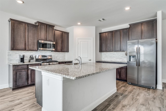 kitchen with sink, appliances with stainless steel finishes, a kitchen island with sink, light stone counters, and light hardwood / wood-style floors