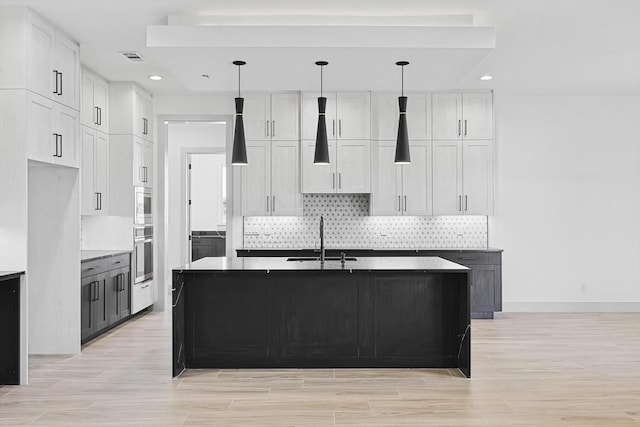 kitchen with stainless steel oven, sink, white cabinetry, and an island with sink