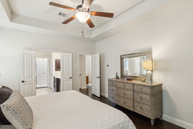 bedroom with a raised ceiling, ceiling fan, dark wood-type flooring, and ensuite bath