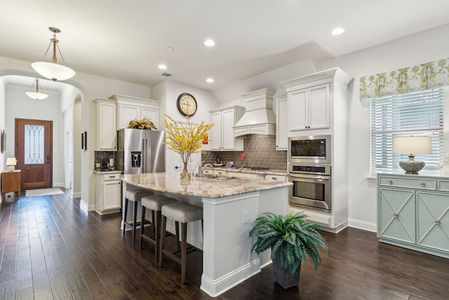 kitchen with appliances with stainless steel finishes, pendant lighting, white cabinetry, custom exhaust hood, and a kitchen island with sink