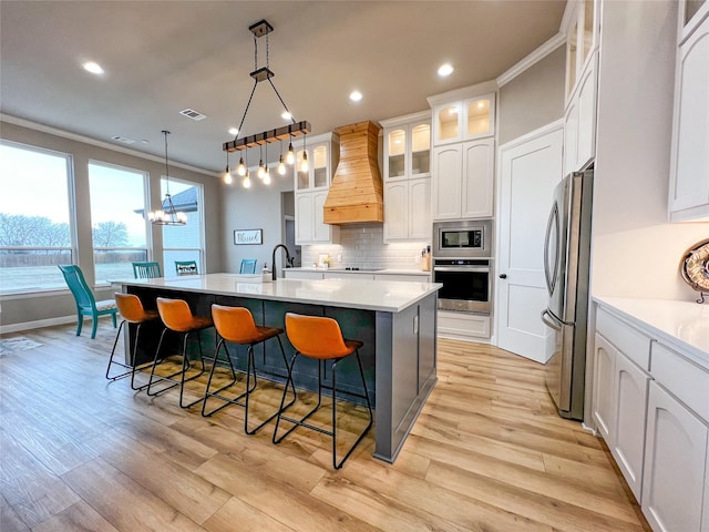 kitchen with a kitchen island with sink, light countertops, appliances with stainless steel finishes, glass insert cabinets, and custom range hood