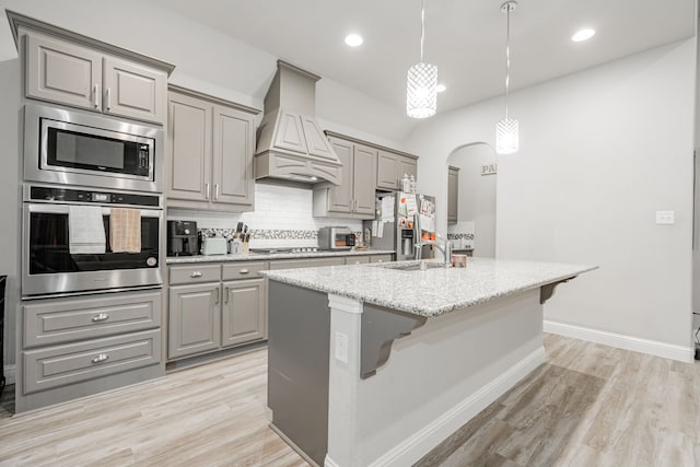 kitchen with light stone counters, custom range hood, stainless steel appliances, a kitchen island with sink, and sink