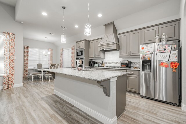 kitchen featuring backsplash, premium range hood, a kitchen island with sink, light stone counters, and stainless steel appliances