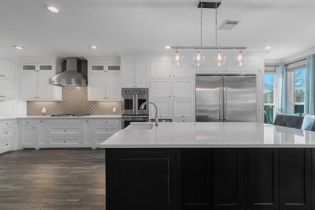 kitchen featuring stainless steel appliances, a kitchen island with sink, hanging light fixtures, and wall chimney exhaust hood