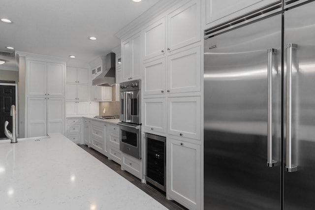 kitchen featuring stainless steel appliances, white cabinets, light stone counters, wall chimney exhaust hood, and beverage cooler