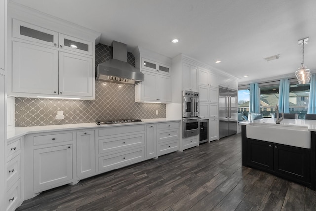 kitchen featuring stainless steel appliances, white cabinetry, wall chimney exhaust hood, hanging light fixtures, and dark wood-type flooring