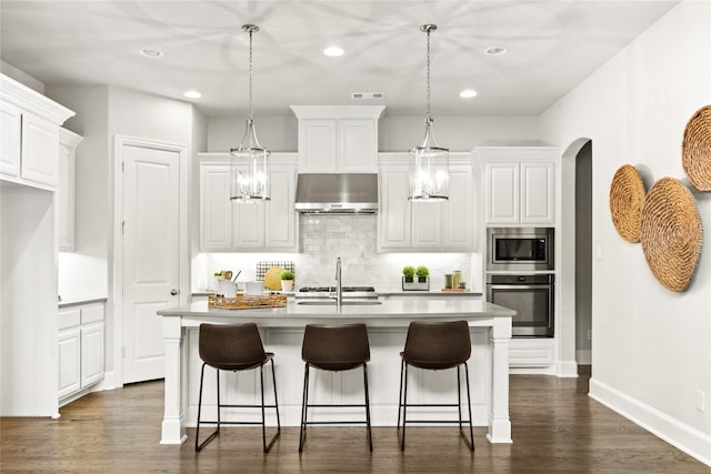 kitchen featuring an island with sink, pendant lighting, wall chimney range hood, and appliances with stainless steel finishes