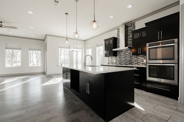 kitchen featuring sink, decorative light fixtures, wall chimney exhaust hood, stainless steel double oven, and a kitchen island with sink