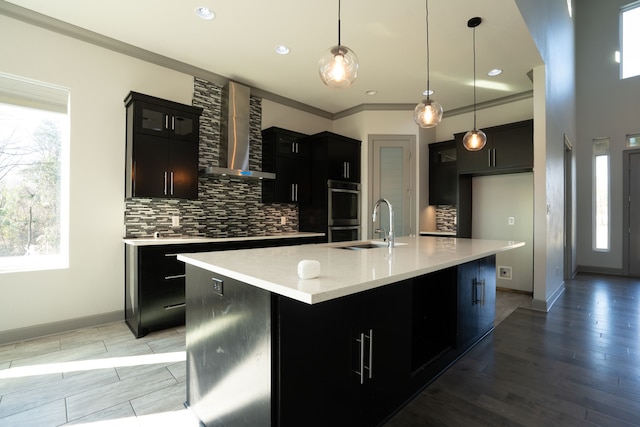 kitchen with hanging light fixtures, an island with sink, crown molding, double oven, and wall chimney exhaust hood