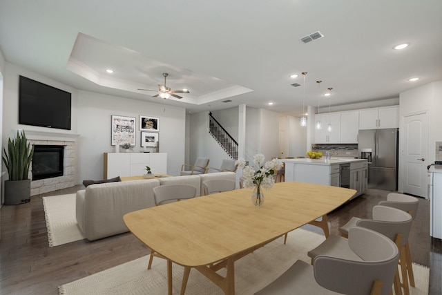 dining room with a tray ceiling, a stone fireplace, ceiling fan, and dark wood-type flooring