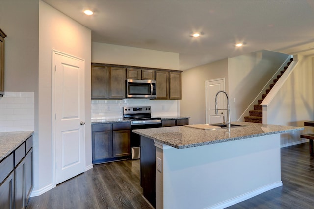 kitchen featuring appliances with stainless steel finishes, tasteful backsplash, light stone counters, a kitchen island with sink, and dark wood-type flooring