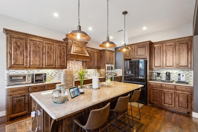 kitchen featuring light stone countertops, appliances with stainless steel finishes, decorative light fixtures, dark wood-type flooring, and a kitchen island with sink