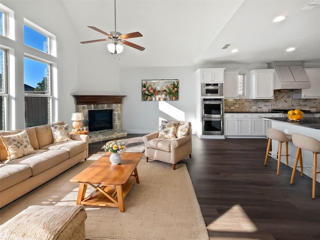 living room with a stone fireplace, dark wood-type flooring, ceiling fan, and a towering ceiling