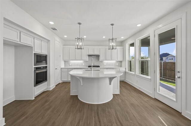 kitchen featuring white cabinetry, hanging light fixtures, an island with sink, built in microwave, and stainless steel oven