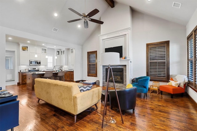 living room featuring a stone fireplace, ceiling fan, beamed ceiling, and dark wood-type flooring