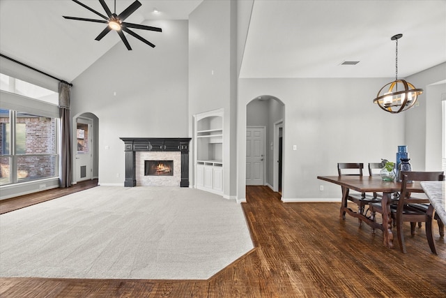 living room featuring high vaulted ceiling, ceiling fan with notable chandelier, a stone fireplace, built in shelves, and dark hardwood / wood-style floors