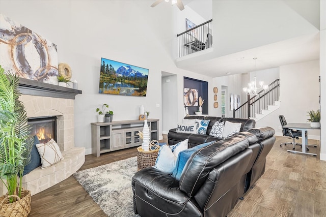 living area featuring stairway, wood finished floors, baseboards, a stone fireplace, and ceiling fan with notable chandelier