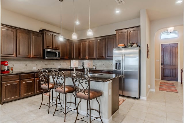 kitchen with dark brown cabinetry, stainless steel appliances, dark stone countertops, pendant lighting, and a kitchen island with sink