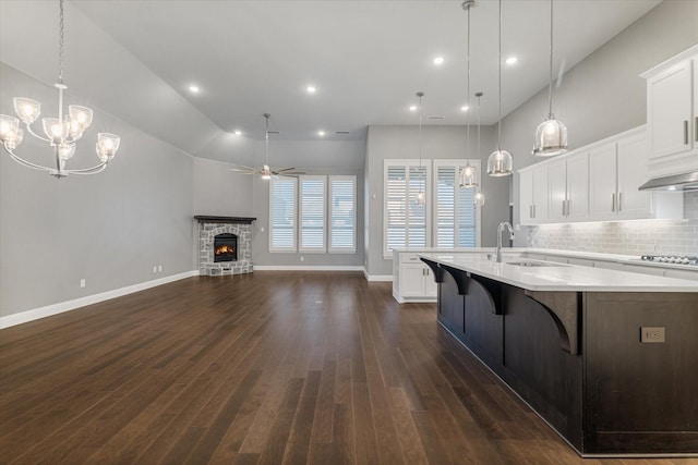 kitchen featuring a kitchen island with sink, light countertops, ceiling fan with notable chandelier, a stone fireplace, and a sink