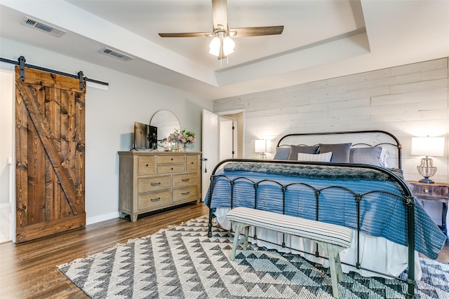 bedroom with a raised ceiling, ceiling fan, a barn door, hardwood / wood-style floors, and wood walls