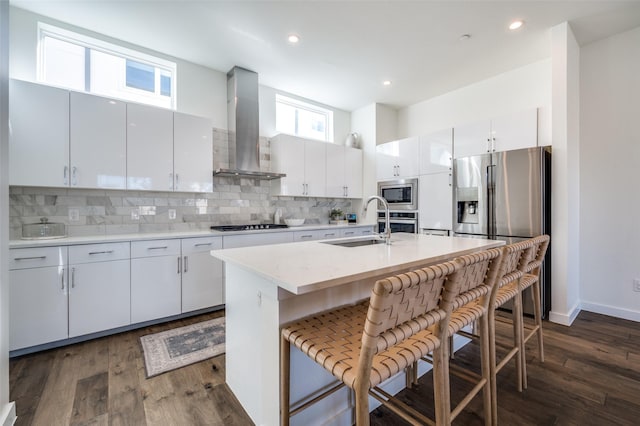 kitchen with stainless steel appliances, white cabinetry, a center island with sink, and wall chimney range hood