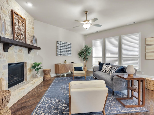 living room with ceiling fan, a stone fireplace, and dark wood-type flooring