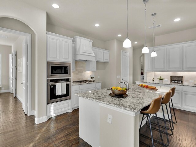 kitchen featuring premium range hood, stainless steel appliances, white cabinets, hanging light fixtures, and an island with sink